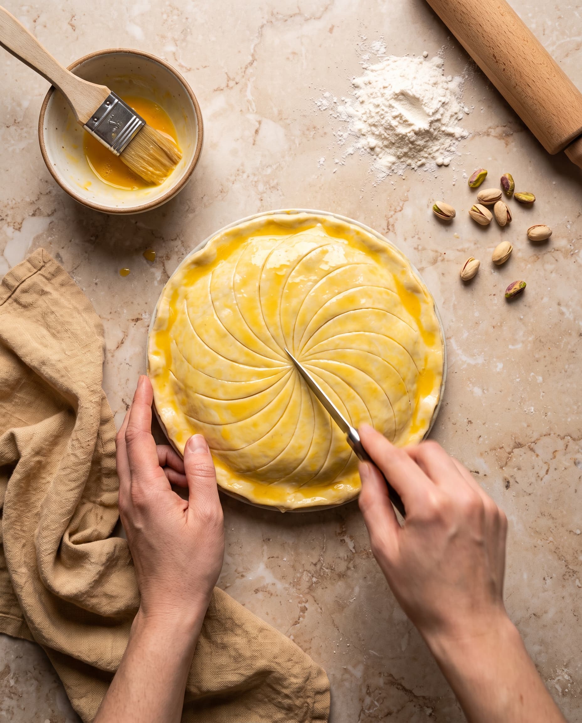 Galette des rois pistache crue en cours de décoration, motif rosace tracé au couteau, dorure à l'œuf au pinceau, mains en action
